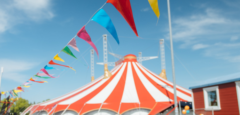 Eine bunte Wimpelkette hängt unter freiem Himmel bei Sonnenschein auf dem Circus Fantasia Platz. Im Hintergrund stehen Menschen und das große Circuszelt.