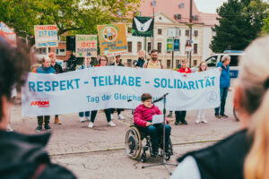 Eine Person im Rollstuhl hält eine Rede, dahinter ist ein Transpi mit der Aufschrift "Respekt - Teilhabe - Solidarität" zu sehen