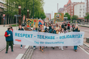 Viele Menschen laufen, rollen und stolzieren hinter dem Transparent mit der Aufschrift "Respekt - Teilhabe - Solidarität, Tag der Gleichstellung"