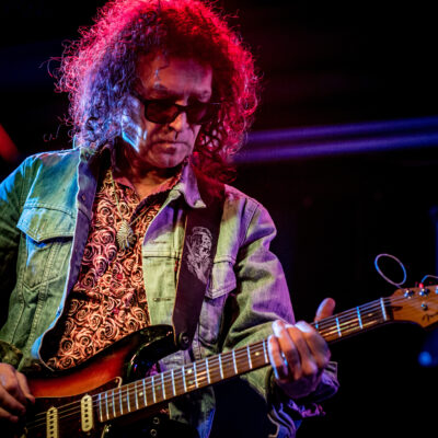 Ein Mann mit langem, lockigem Haar und Sonnenbrille, spielt in einem Jeanshemd gekleidet eine E-Gitarre